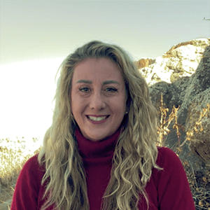 A smiling woman with long curly blonde hair wearing a red turtleneck, standing outdoors with sunlit hills in the background.