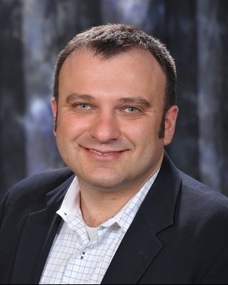 A portrait of a smiling man with short hair, wearing a dark suit jacket over a white shirt, against a mottled gray backdrop.