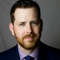 A professional headshot of a man with a beard, wearing a suit and tie, looking directly at the camera. He has short brown hair and a confident expression.