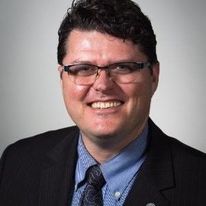 Headshot of a smiling man with glasses, wearing a dark suit, blue patterned tie, and a blue striped shirt against a gray background.
