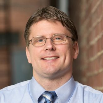 A smiling man with glasses, wearing a light blue shirt and striped tie, stands in front of a brick wall background.