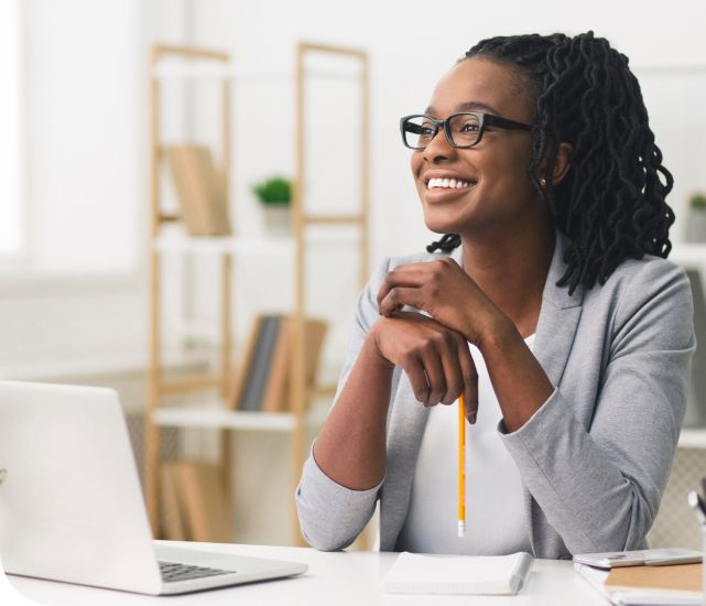 A joyful black woman with glasses and braided hair, wearing a grey blazer, sits at a desk with a laptop and notebook, looking up and away with a thoughtful smile. She is contemplating her future as she applies for scholarships for master's degree students.