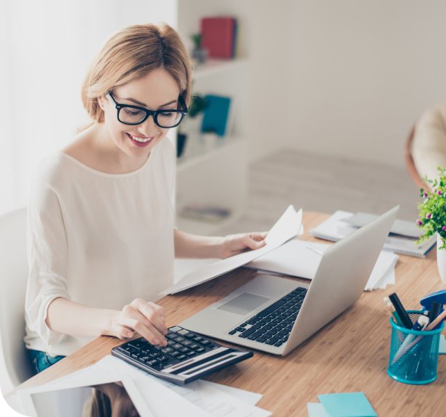 A smiling woman with glasses works at a desk, using a laptop and calculator to manage her financial aid for an online master's degree, surrounded by papers and office supplies.