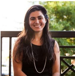 A smiling woman with long dark hair, wearing a black top and a pearl necklace, seated outdoors with a blurred green background.