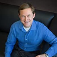 A man in a blue shirt smiling and sitting on a black sofa in a well-lit room. He appears relaxed and is looking directly at the camera.