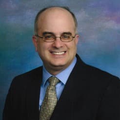 A professional portrait of a smiling bald man wearing glasses, a dark suit, blue shirt, and patterned tie, against a softly blurred multicolored background.