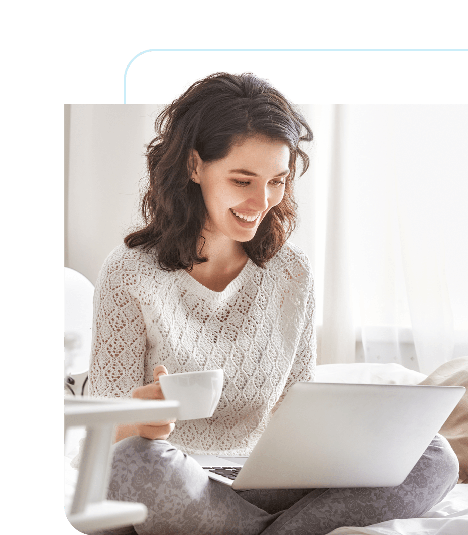 A smiling woman in a white sweater sits comfortably with her laptop and a coffee cup, enjoying a sunny day indoors.