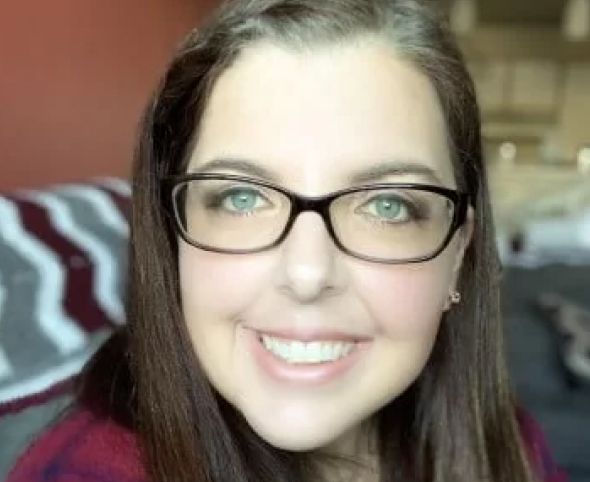 A close-up portrait of a smiling woman with glasses, featuring light skin, dark hair, and striking green eyes. She is inside a room with soft, natural lighting.