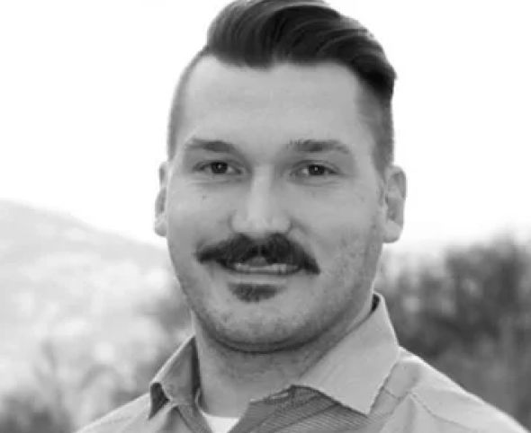 About us: Black and white portrait of a cheerful man with a styled mustache, wearing a collared shirt, and a blurred natural background.