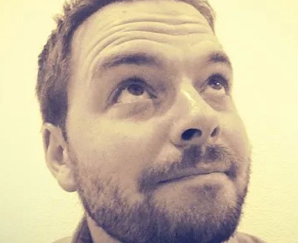 A sepia-toned close-up photo of a Caucasian man looking upwards with a thoughtful expression and a slight smile, showcasing his stubble and bright eyes.