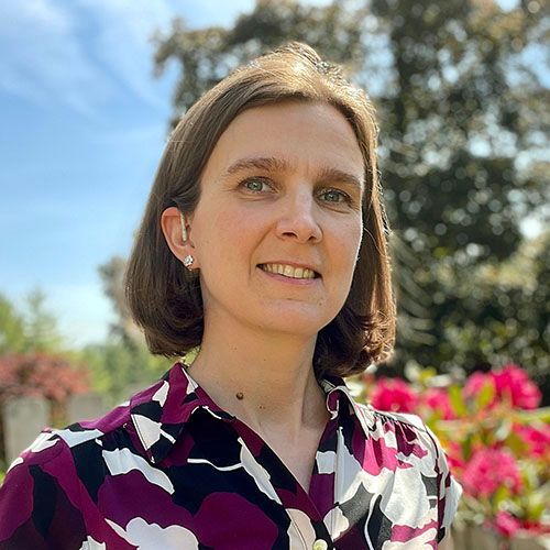 A woman with short brown hair, smiling, wearing a black and white patterned blouse, standing outdoors with sunlight and blurred green foliage and pink flowers in the background. She is a student in an online master's program.