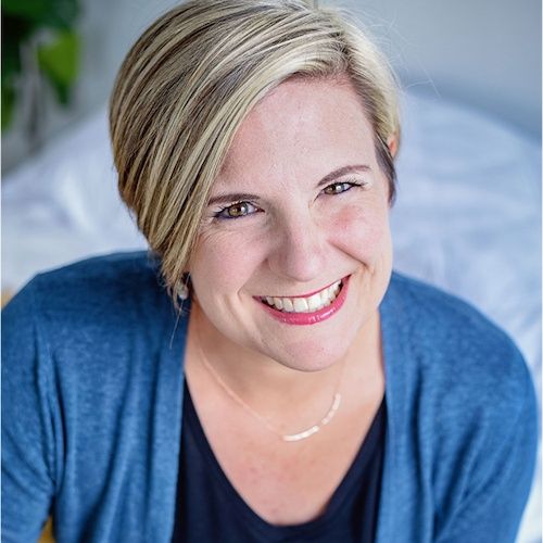 A smiling woman with short blonde hair wearing a blue cardigan over a white top, with a subtle necklace, photographed from above against a blurred background.