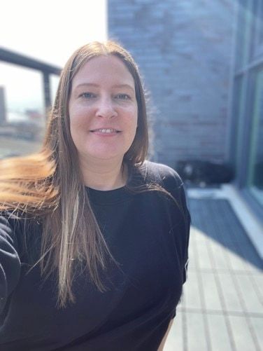 A woman with long brown hair in a black top smiles while taking a selfie outdoors, with a blurred background featuring modern architecture.