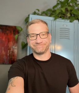 A smiling man with glasses and a beard, wearing a black t-shirt, stands in a room with a locker and plants in the background.