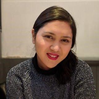 A woman smiling gently at the camera, wearing a gray knitted sweater and sitting indoors. She has shoulder-length dark hair and is wearing red lipstick.