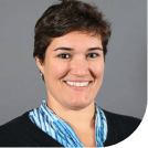 A professional headshot of a smiling woman with short hair, wearing a black jacket and a blue striped scarf. The background is grey.