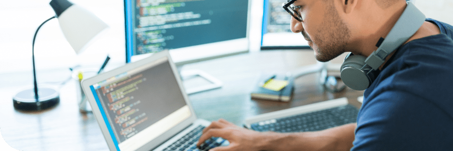 A man with headphones around his neck is coding on a laptop in a well-lit office, with a second monitor displaying code and a desk lamp turned on.