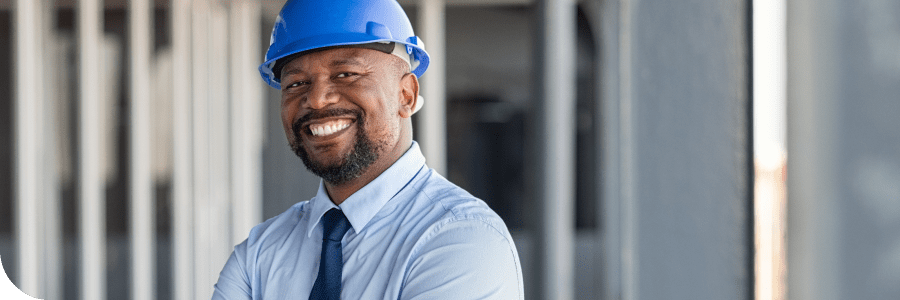 A smiling man with a beard, wearing a blue hard hat and a light blue shirt, standing confidently at a construction site.