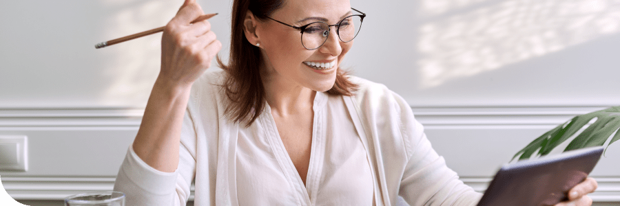 A smiling woman with glasses, holding a pencil and a notebook, looks at the notes she is making. She appears to be sitting in a bright room with sunlight.