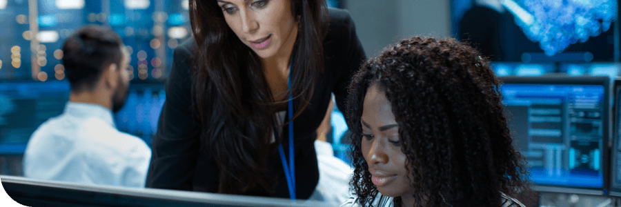 Two professional women, one standing and one seated, analyze data on a computer screen in a high-tech office environment. Other colleagues visible in the background.