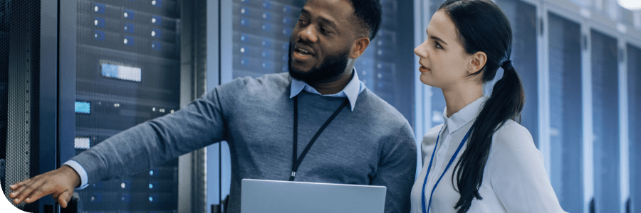 Two professionals, a man and a woman, are analyzing data in a server room, with the man pointing at a server rack and the woman holding a laptop.