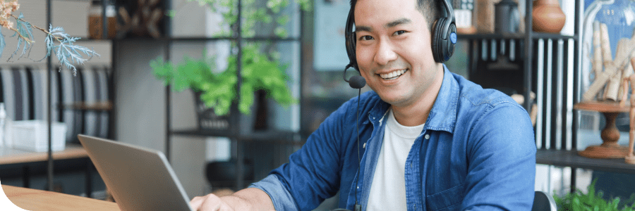 A smiling Asian man wearing a headset and working on a laptop in a modern office setting.