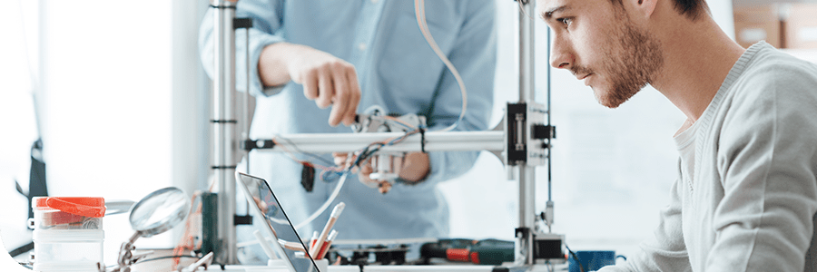 Two men working intently on a 3D printer in a tech workshop, surrounded by various tools and electronic devices.