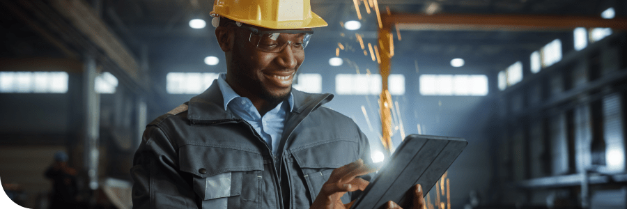 A smiling African-American man wearing a safety helmet and goggles, using a tablet in an industrial setting with sparks flying in the background.