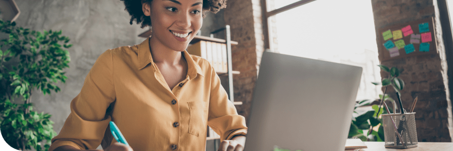 A smiling young woman in a yellow blouse working on a laptop at her desk with sticky notes on the wall in the background.