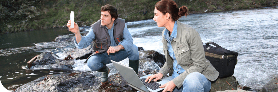 A man and a woman by a riverbank; the man takes a photo with his smartphone while the woman works on a laptop, both wearing casual outfits.