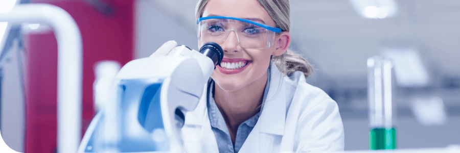 A female scientist wearing safety goggles and a lab coat smiles while using a microscope in a laboratory setting.