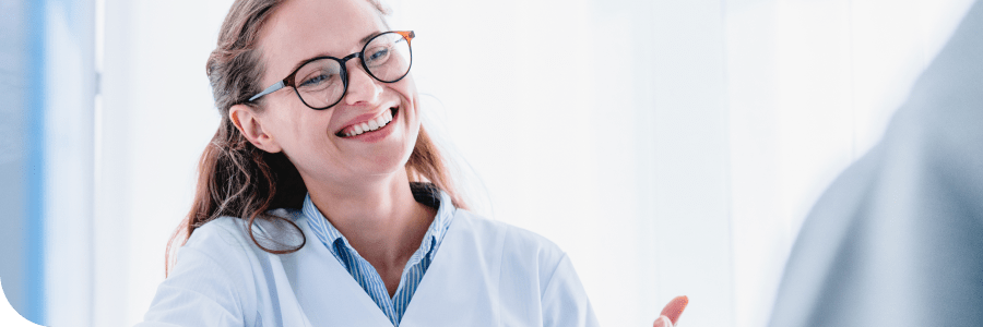 A smiling female doctor with glasses, wearing a lab coat over a blue blouse, interacts with a patient in a bright clinical setting.