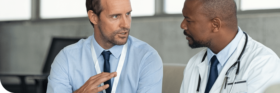 Two male doctors in discussion, one Caucasian and one African American, wearing white coats and blue shirts in a hospital office.