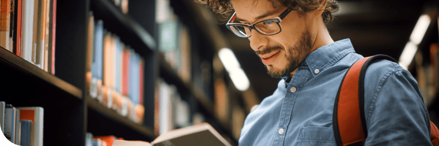 A young man with glasses and a beard, wearing a denim shirt and a backpack, is reading a book in a library, surrounded by shelves filled with books.