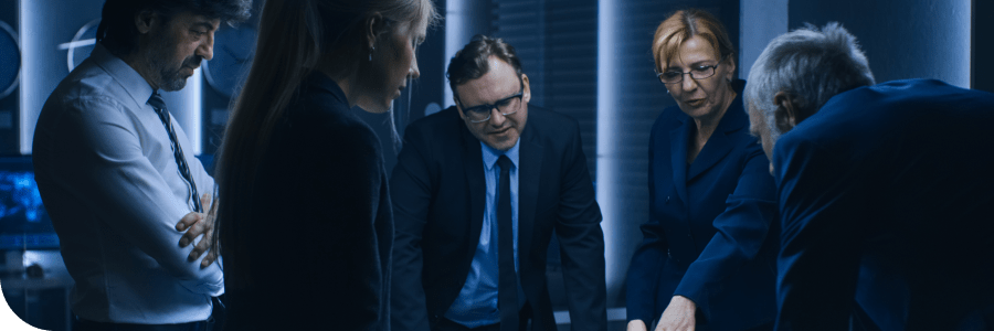 Five professionals, wearing business attire, discuss intently in a dimly lit office environment, evoking a serious, strategic atmosphere.