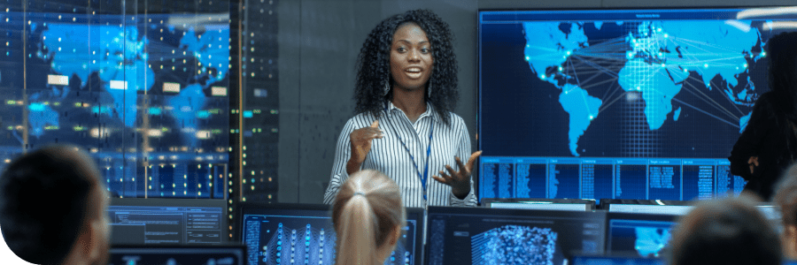 A confident Black woman presents in a high-tech command center with multiple screens displaying data and a world map, addressing an attentive team.