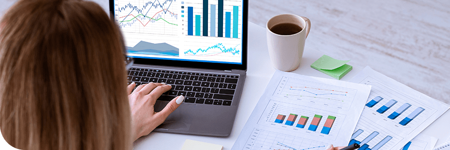 A woman analyzes financial graphs for her Online Economics MBA program on her laptop and printed charts at a white desk, with a coffee cup beside her.
