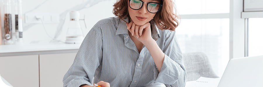 A woman with glasses in a striped shirt intently works on an Online Finance MBA program on her laptop in a bright, modern office, with a thoughtful expression while holding a yellow pencil.