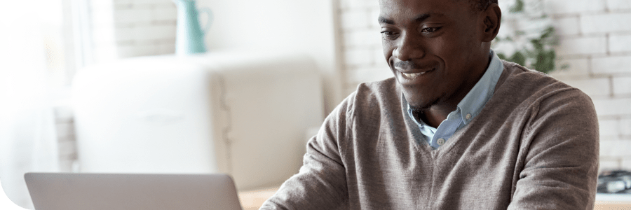 A man smiling while working on a laptop in a bright, well-lit room, suggesting a comfortable and productive environment for engaging with the best online MBA programs in 2023.