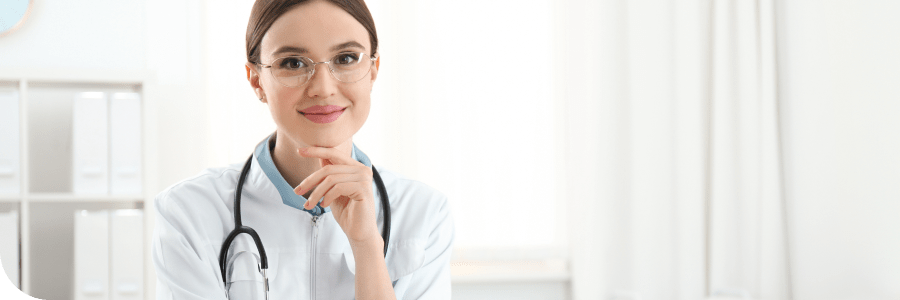 A female doctor in a white coat with a stethoscope around her neck, smiling and touching her chin, sitting at a desk in a bright office.