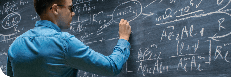 A man in a blue shirt writes complex mathematical equations on a large blackboard filled with formulas.