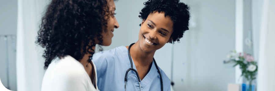 A smiling female nurse with a stethoscope talks to a female patient in a medical office, conveying warmth and professionalism.