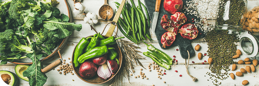 A variety of fresh vegetables and fruits spread on a wooden surface, including spinach, pomegranate, green beans, avocados, and almonds.