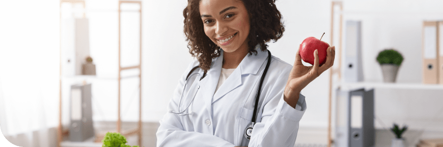 A smiling female doctor in a white coat holding a red apple in a bright, modern office.