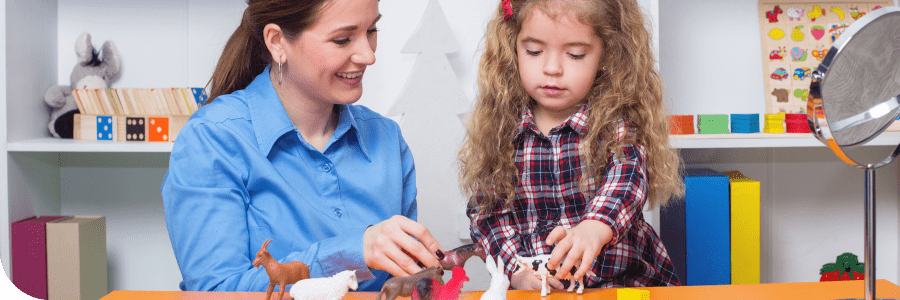 A young girl and a woman, possibly a teacher, engaged in play with toy animals on a colorful table in a classroom setting.