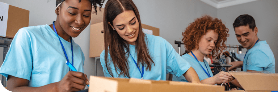 Four young volunteers wearing blue t-shirts are happily packing items into boxes in a room with cardboard boxes around, suggesting a community service activity.