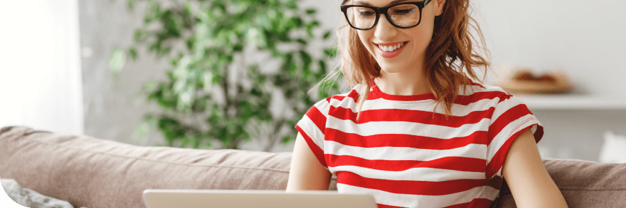 A smiling woman with glasses, wearing a striped red and white shirt, works on a laptop while sitting comfortably on a couch in a bright, plant-decorated room.