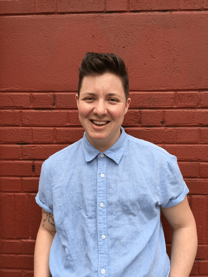 A person with a short haircut smiles in front of a red brick wall, wearing a light blue button-up shirt.