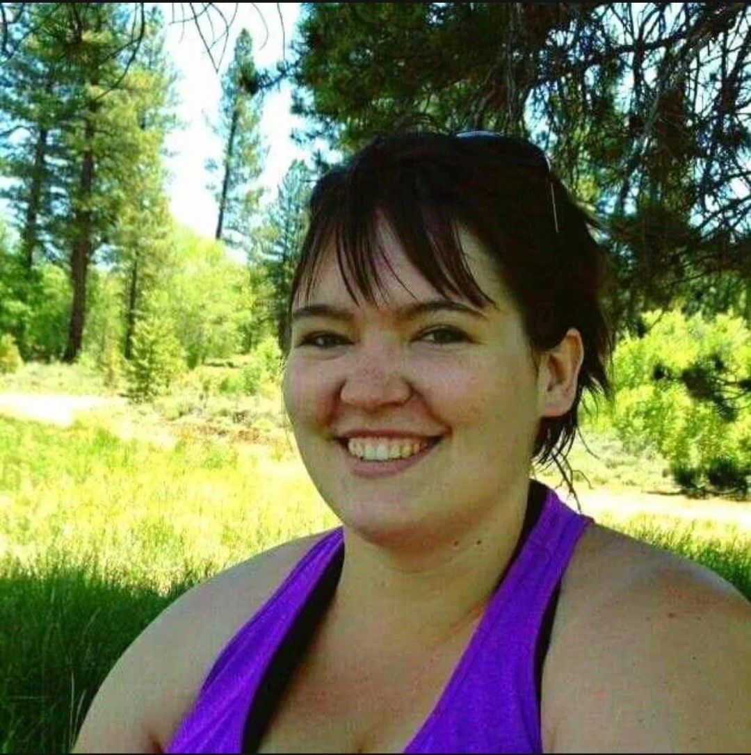 A smiling woman with shoulder-length dark hair, wearing a purple tank top, against a backdrop of pine trees and a grassy area under a bright sky.