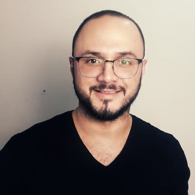 A portrait of a smiling man with a beard, wearing glasses and a black V-neck shirt against a light gray background.